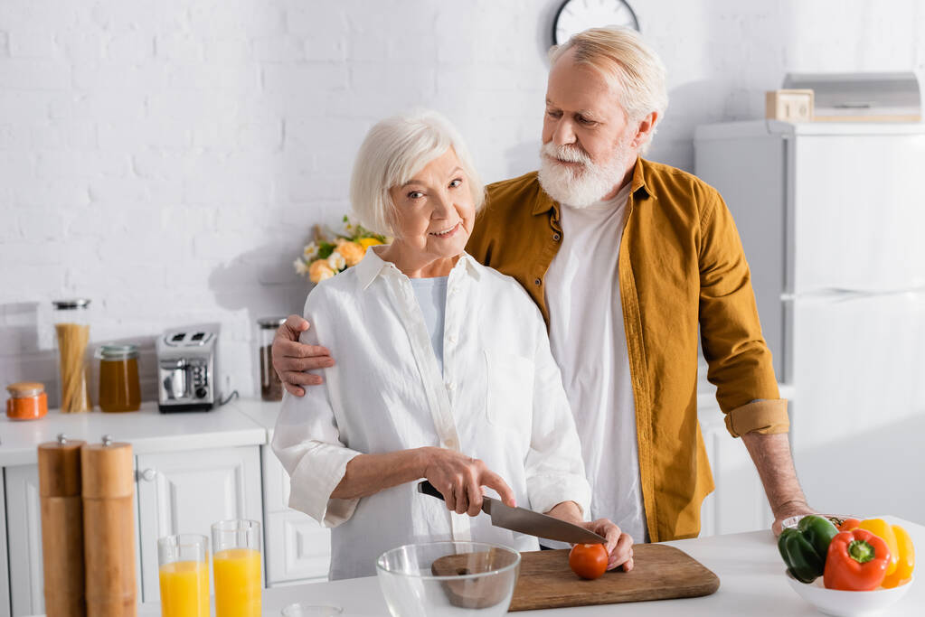 Yaşlı bir adam mutfakta yemek yaparken karısını kucaklıyor domates kesiyor.  - Fotoğraf, Görsel