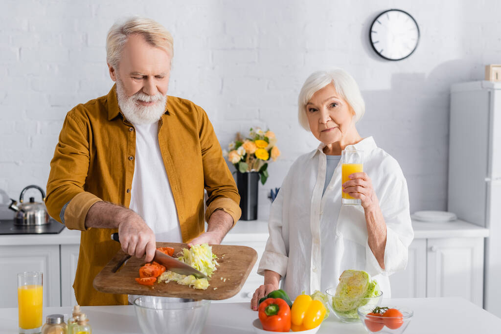 Ηλικιωμένη γυναίκα που κρατά ένα ποτήρι χυμό πορτοκαλιού κοντά στο σύζυγο ρίχνει λαχανικά σε μπολ στην κουζίνα  - Φωτογραφία, εικόνα