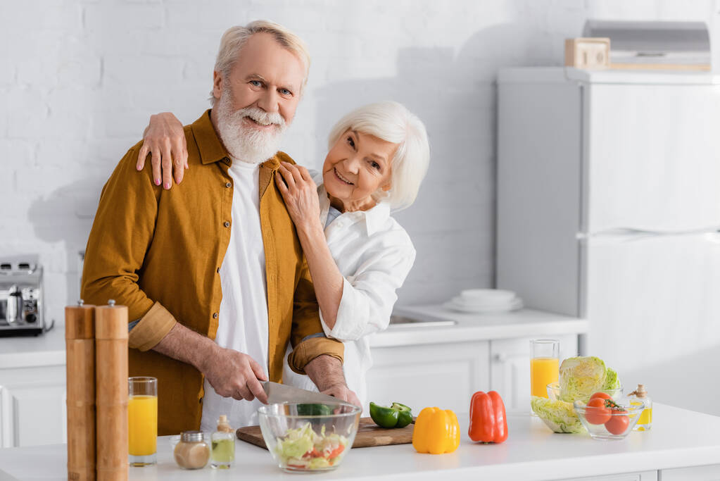 Улыбающаяся женщина обнимает старшего мужа, режет овощи рядом с апельсиновым соком и миской на кухне  - Фото, изображение