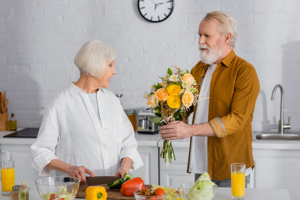 Ανώτερος άνδρας παρουσιάζει λουλούδια στη σύζυγο κοπής λαχανικών στην κουζίνα  - Φωτογραφία, εικόνα