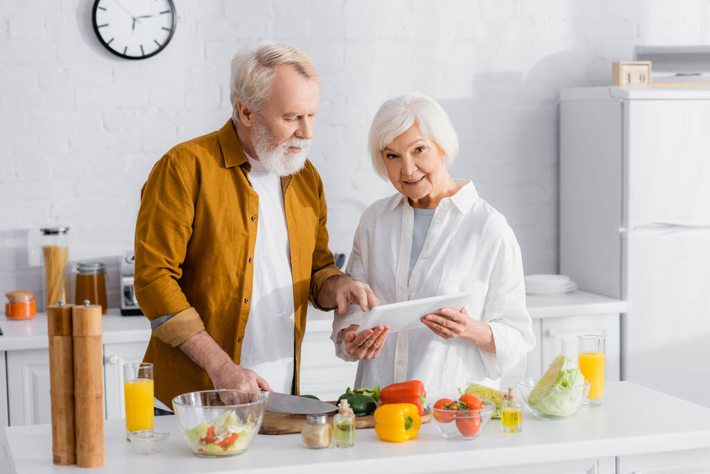 Старша жінка з цифровим планшетом, що стоїть біля чоловіка, який готує на кухні
  - Фото, зображення