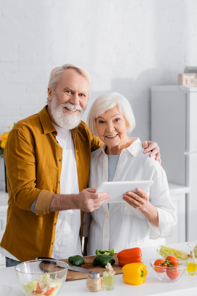 Joyeux couple de personnes âgées tenant tablette numérique près de la planche à découper et légumes au premier plan flou dans la cuisine   - Photo, image