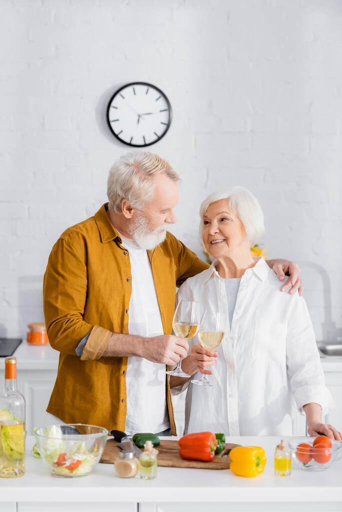 Старший мужчина обнимает жену во время звона с вином возле овощей на размытом переднем плане на кухне  - Фото, изображение