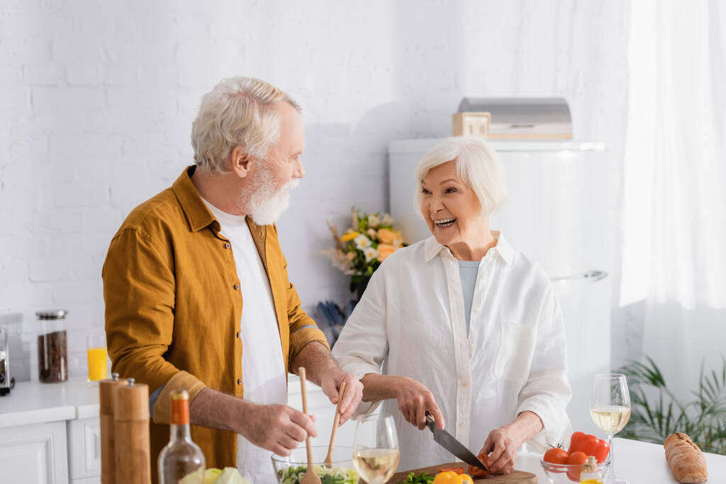 Vidám idős nő vágás zöldségek, míg a férj keverés saláta bor közelében a konyhában  - Fotó, kép