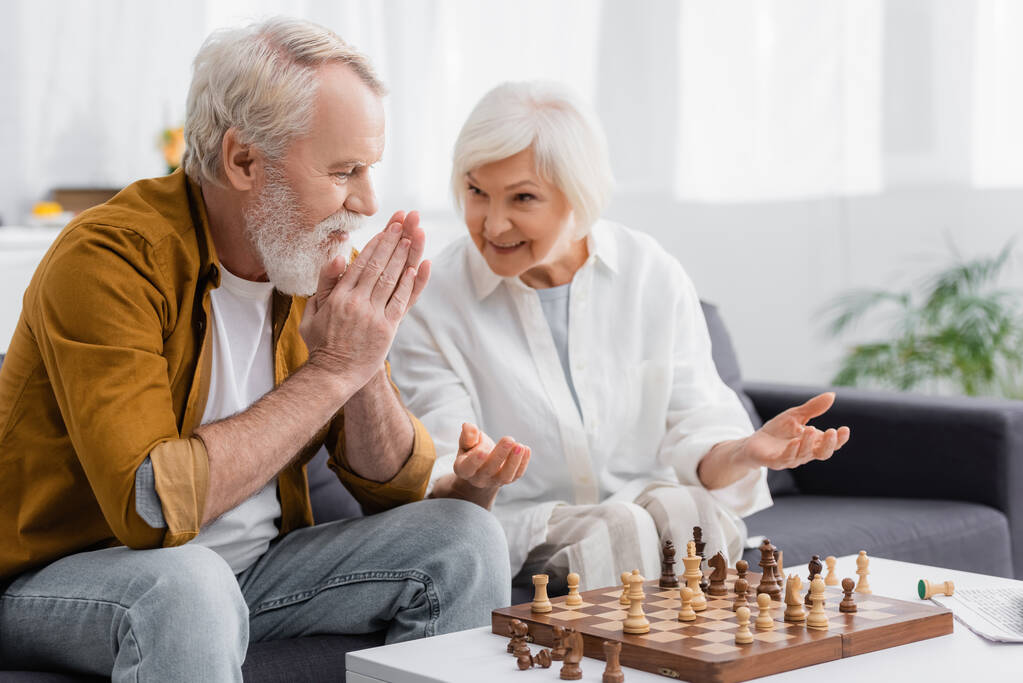 Senior man kijkt naar schaken in de buurt van glimlachende vrouw op wazige achtergrond  - Foto, afbeelding