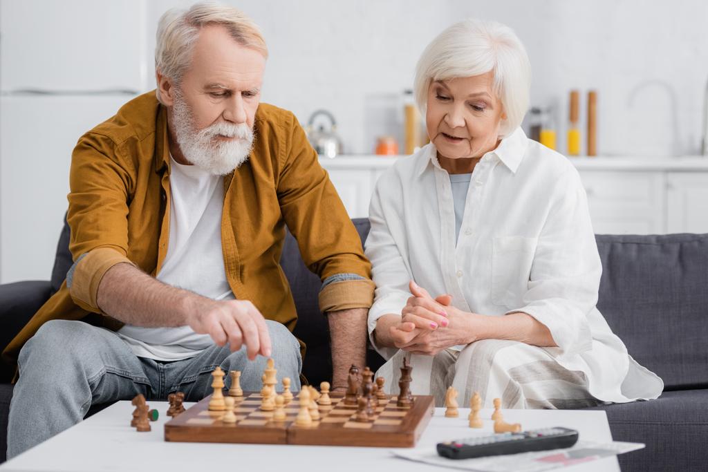 Couple âgé jouant aux échecs près du journal et de la télécommande au premier plan flou  - Photo, image