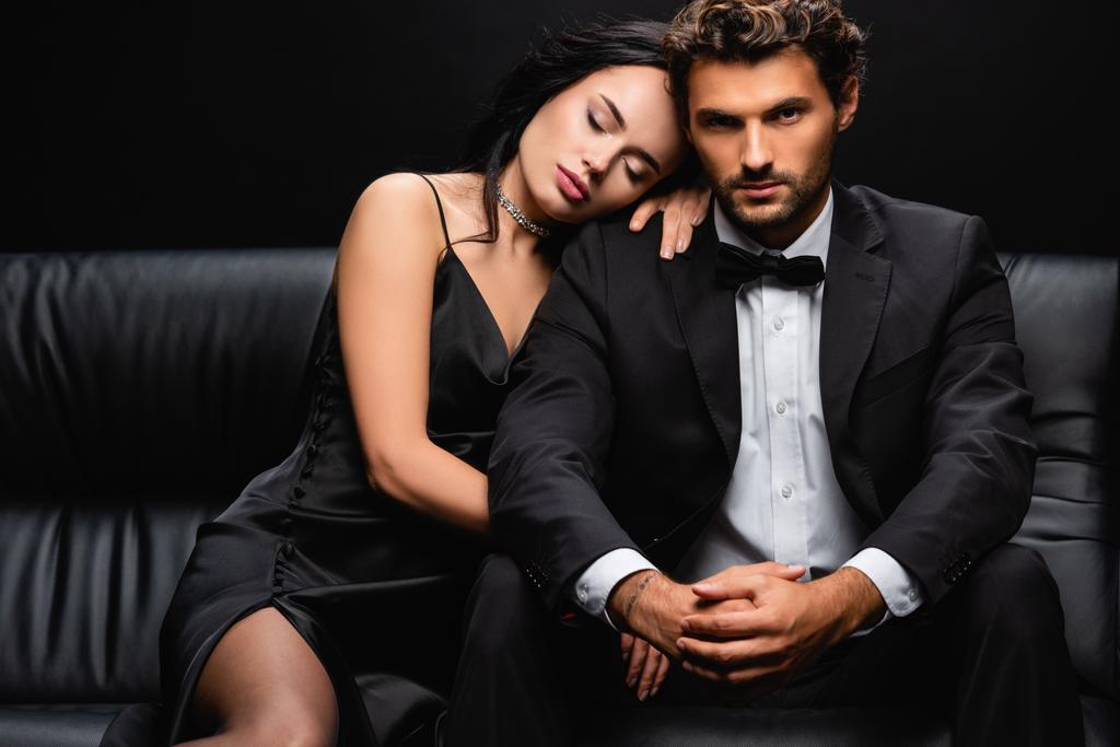 sexy Frau im Satinkleid auf der Schulter eines eleganten Mannes sitzend auf Ledercouch isoliert auf schwarz - Foto, Bild