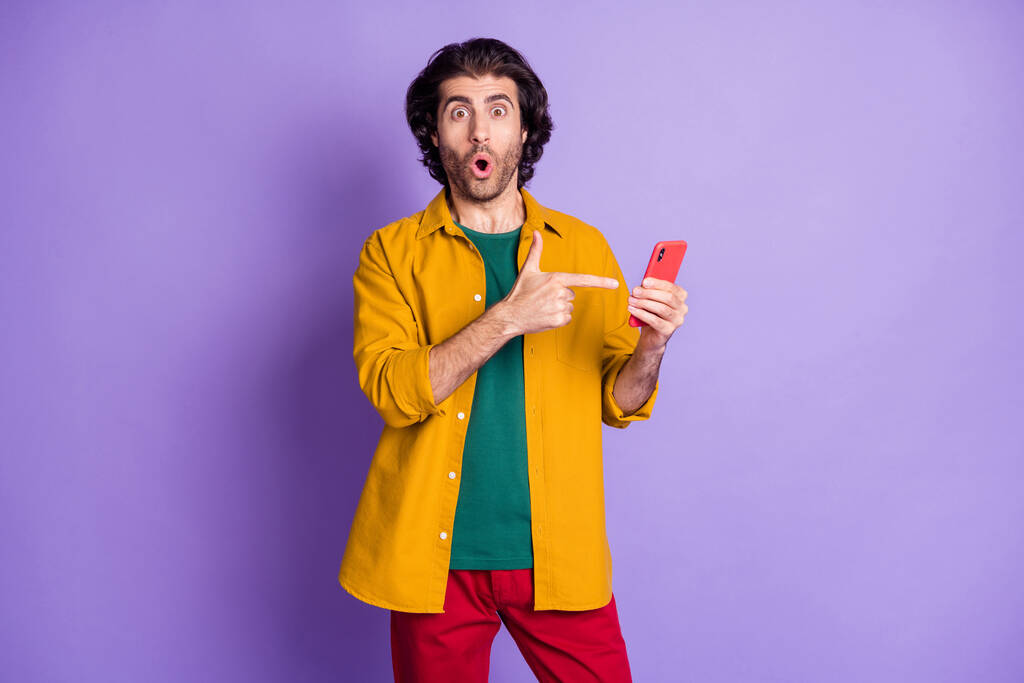 Photo de jeune beau surpris surpris homme homme mâle pointant du doigt sur le téléphone portable isolé sur fond de couleur violette - Photo, image