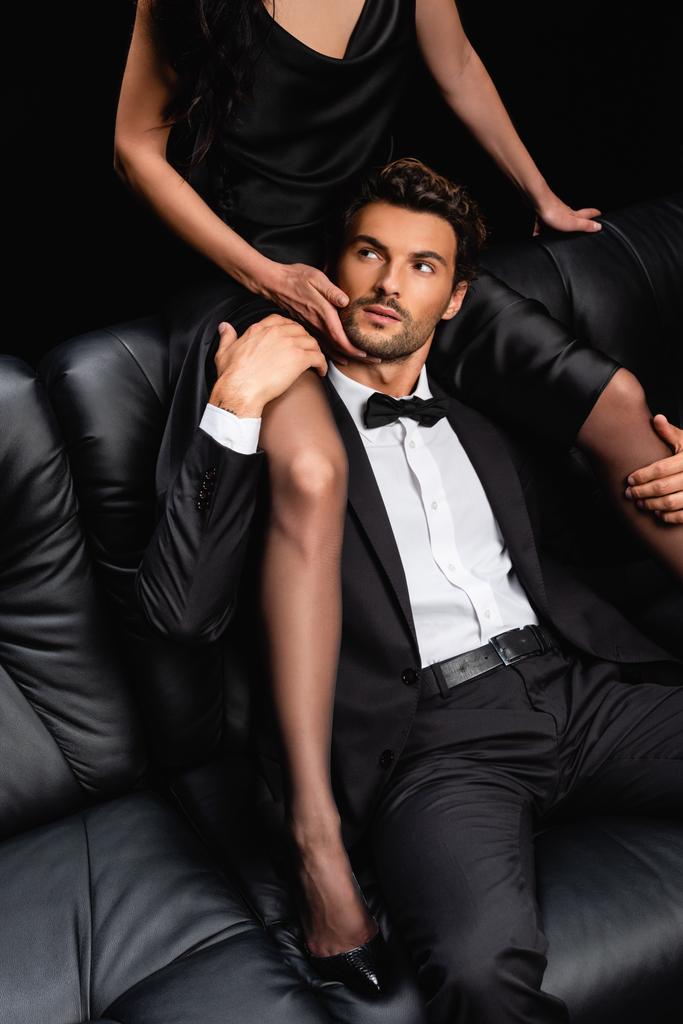 σέξι γυναίκα κάθεται στον καναπέ πάνω από τον άνδρα με κοστούμι και αγγίζοντας το πρόσωπό του απομονωμένο σε μαύρο - Φωτογραφία, εικόνα