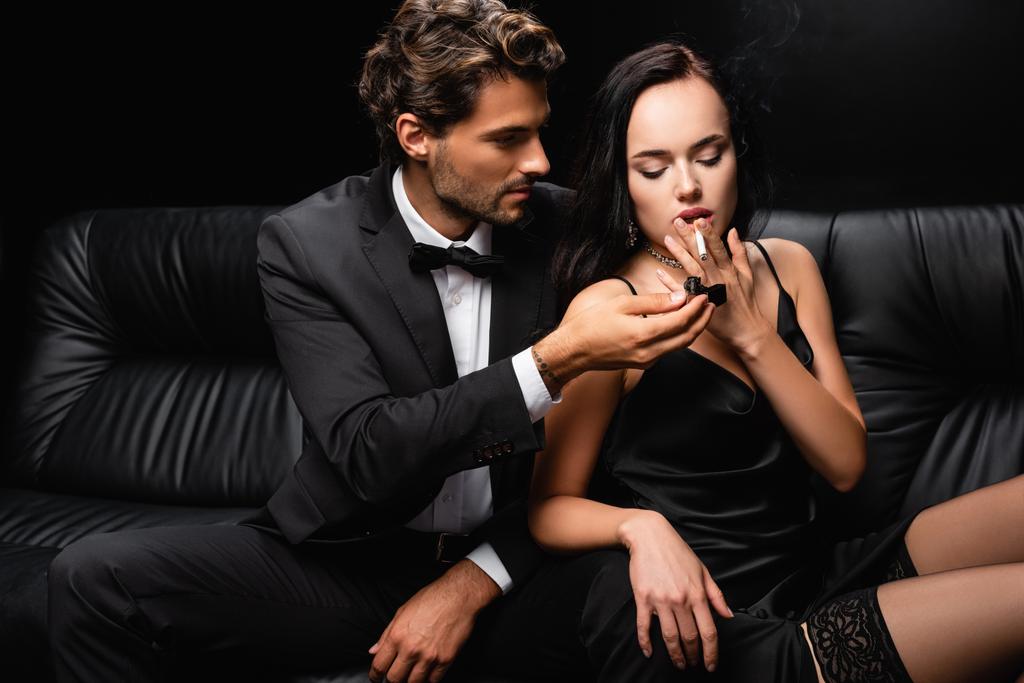 Mann im Anzug blitzt Zigarette der sinnlichen Frau im schwarzen Satinkleid isoliert auf schwarz - Foto, Bild