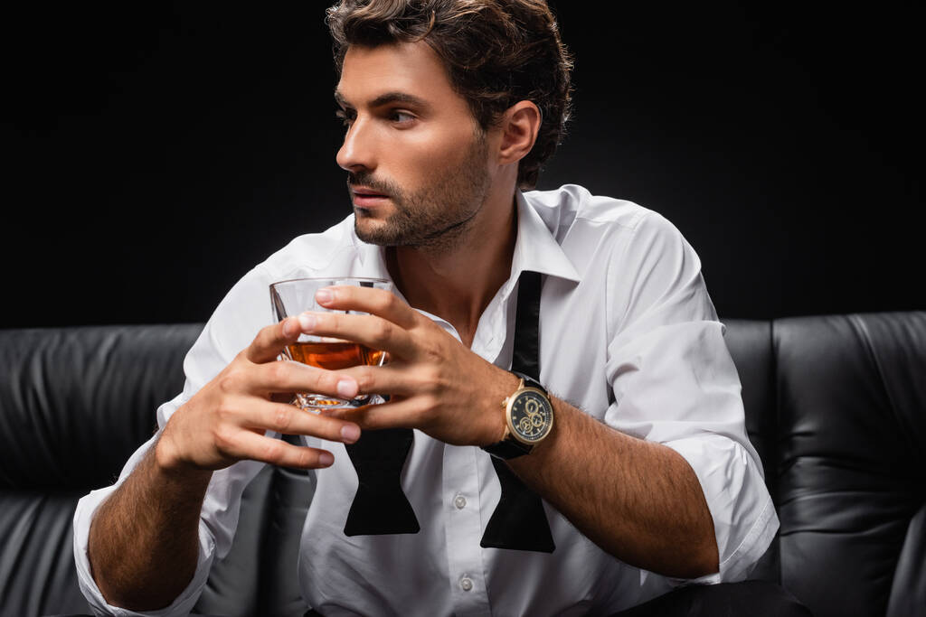 młody człowiek w rozwiązanym krawacie i białej koszuli odwracając wzrok trzymając szklankę whisky odizolowanej na czarno - Zdjęcie, obraz