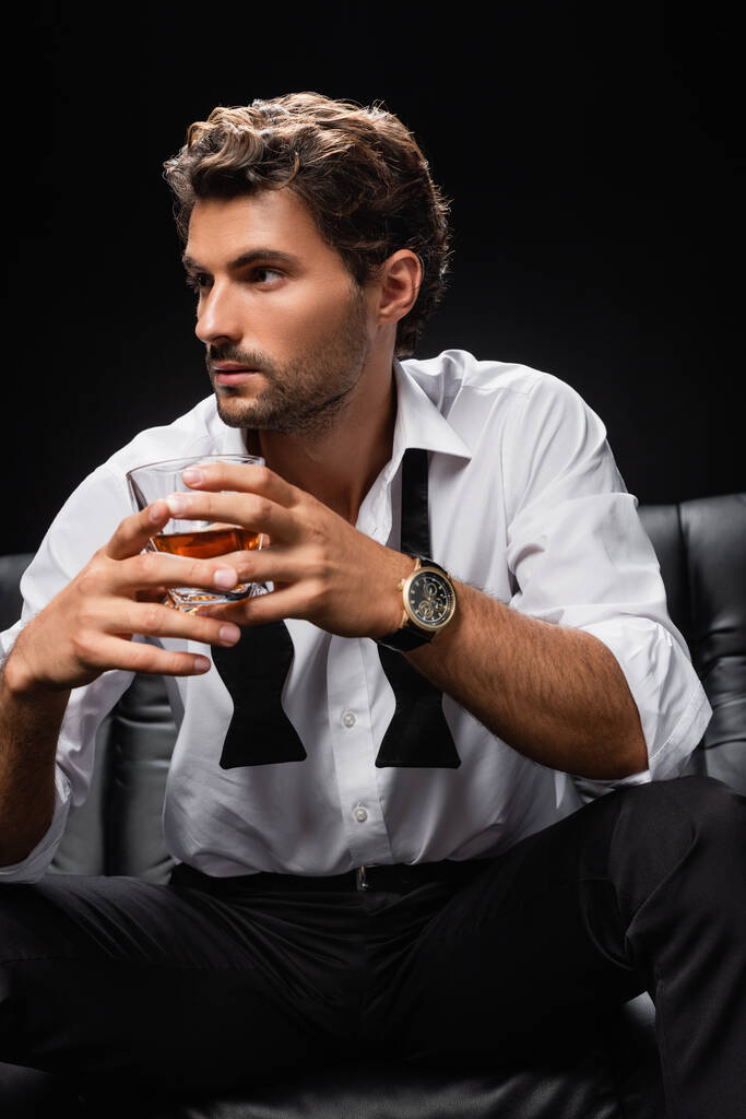 giovane uomo in camicia bianca e cravatta slegata con un bicchiere di whisky mentre guarda lontano isolato sul nero - Foto, immagini