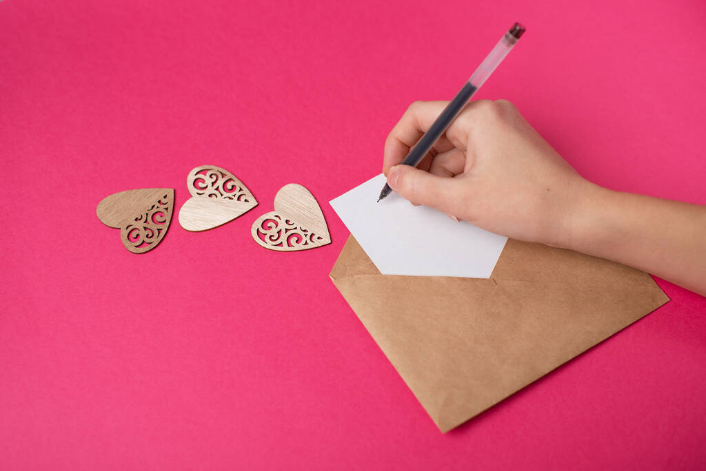 Vrouwenhand die een pen over de ambachtelijke envelop houdt met een blanco vel papier op de hete roze achtergrond. - Foto, afbeelding