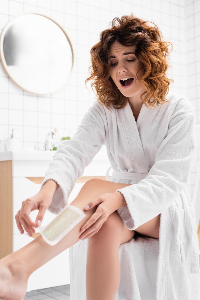 Sikoltozó nő fürdőköpenyben eltávolítja a szőrtelenítő csíkot a lábáról a fürdőszobában  - Fotó, kép
