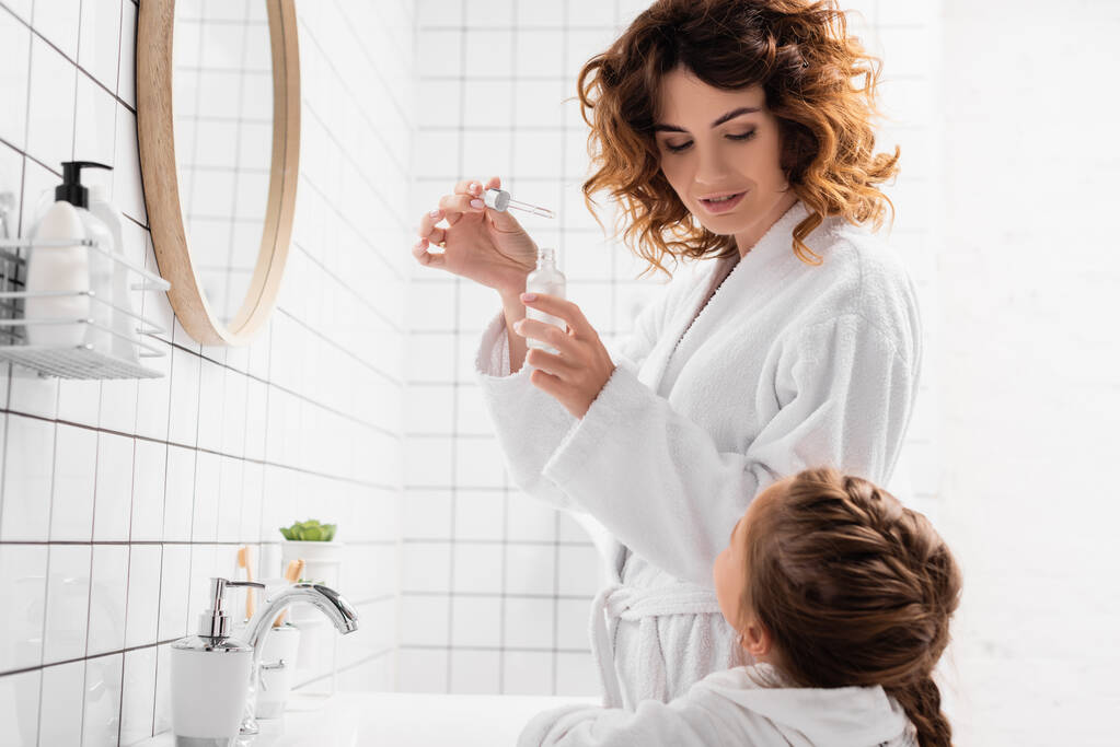 Женщина в халате с косметической сывороткой рядом с ребенком в ванной комнате  - Фото, изображение