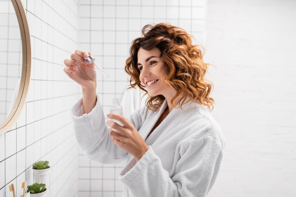 Femme souriante en peignoir tenant sérum cosmétique dans la salle de bain  - Photo, image