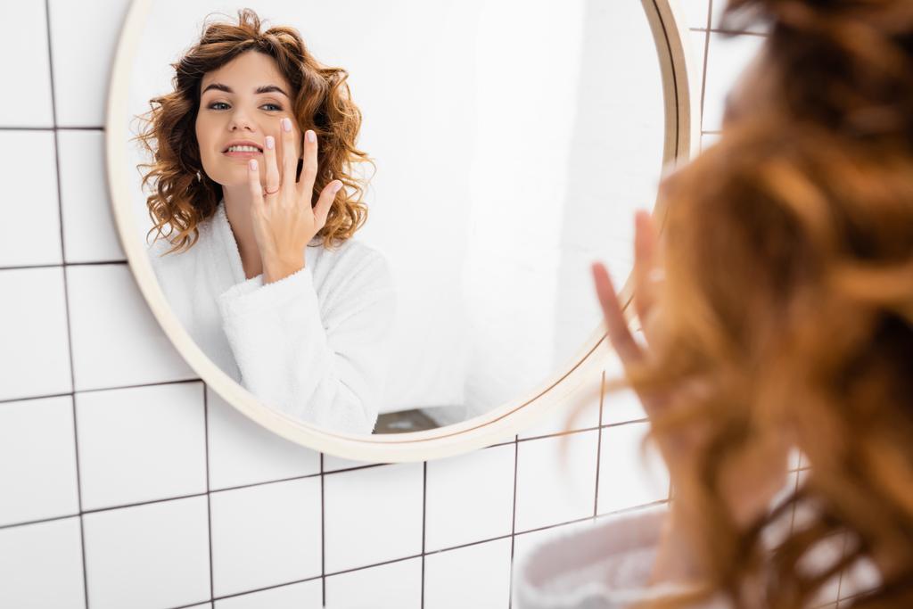 Mulher sorridente em roupão aplicando creme facial perto do espelho em primeiro plano borrado  - Foto, Imagem
