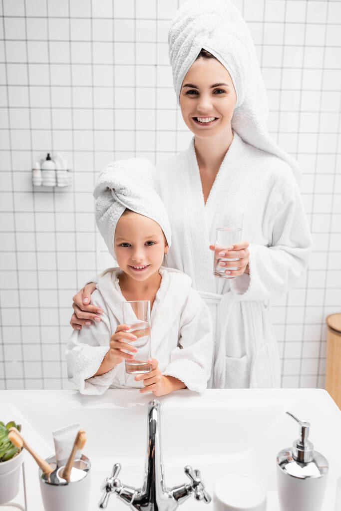 Sonrientes madre e hija en albornoces y toallas sosteniendo vasos de agua en el baño  - Foto, imagen
