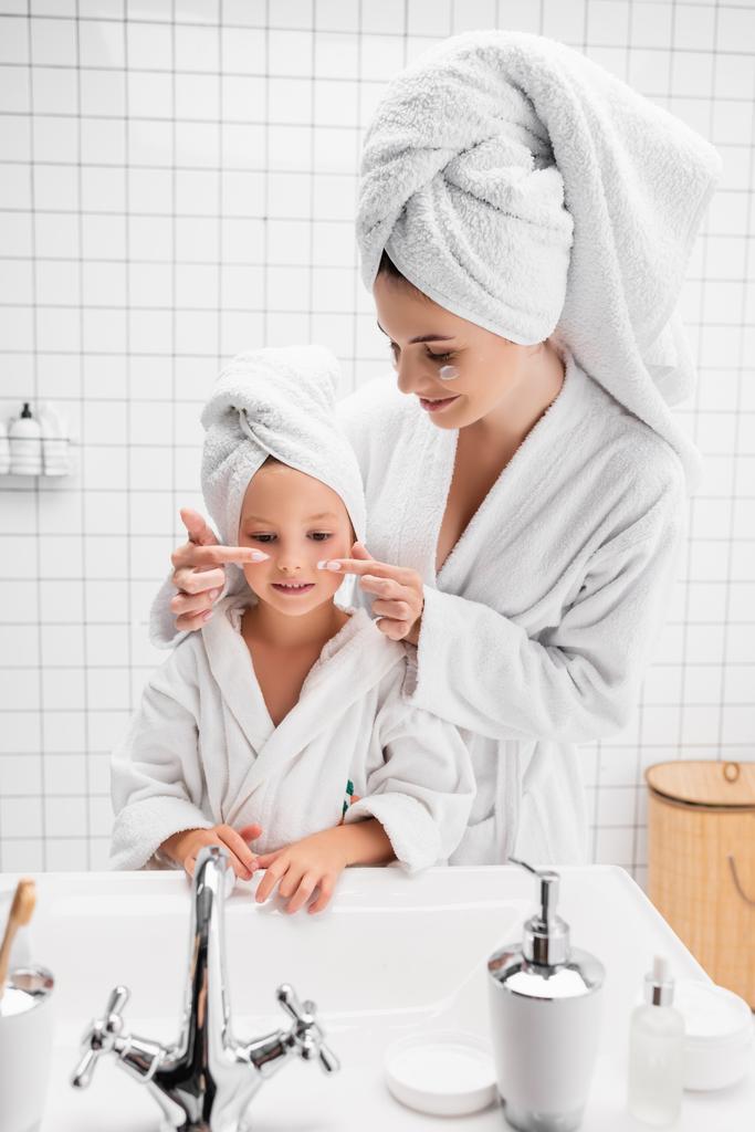 Moeder aanbrengen gezicht crème op dochter wangen in de badkamer  - Foto, afbeelding