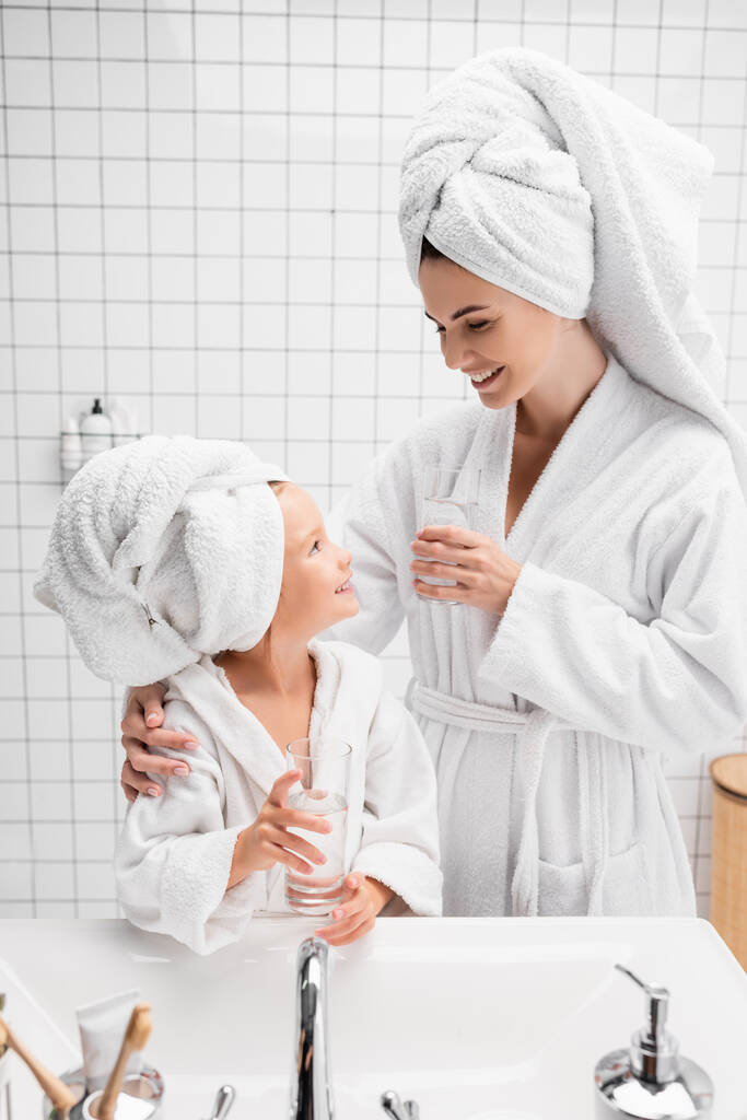Χαμογελαστή μητέρα με μπουρνούζι και πετσέτα κρατώντας ένα ποτήρι νερό κοντά στην κόρη της στο μπάνιο  - Φωτογραφία, εικόνα
