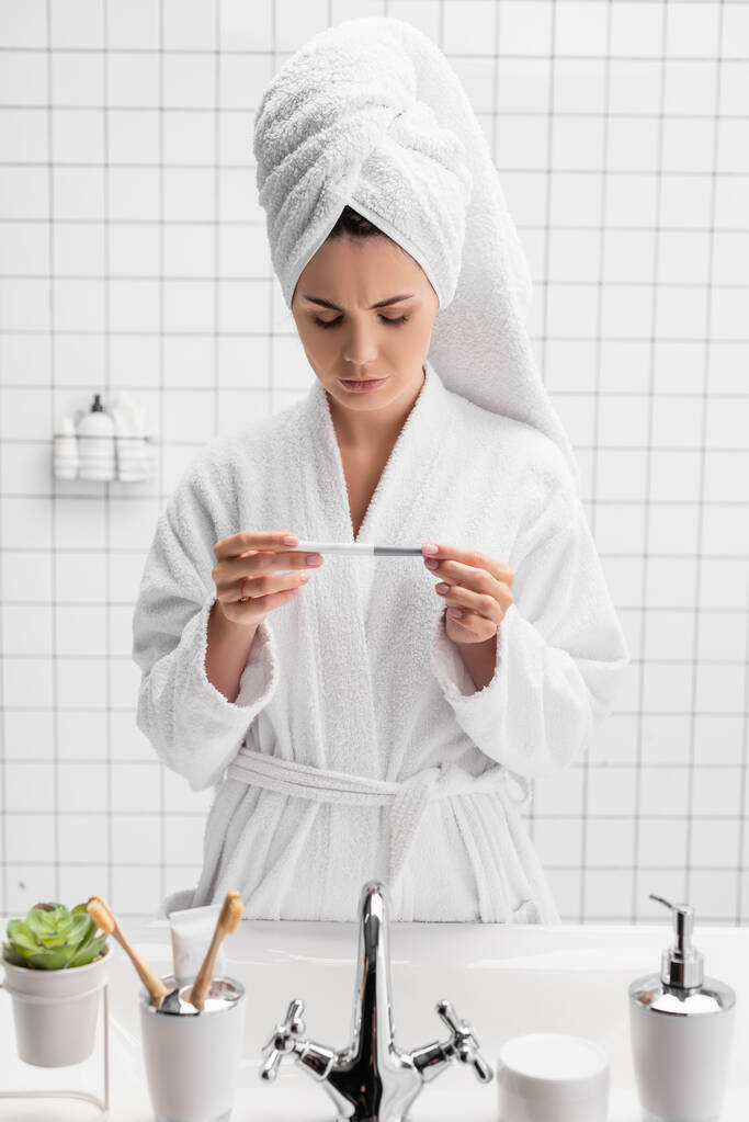 Triste femme en serviette et peignoir tenant test de grossesse dans la salle de bain  - Photo, image