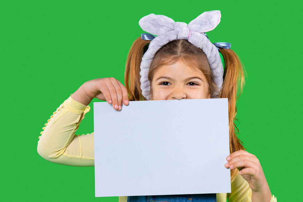 Дошкільна дівчина з пов'язкою на голові тримає білий лист для вашої реклами на зеленому тлі
. - Фото, зображення