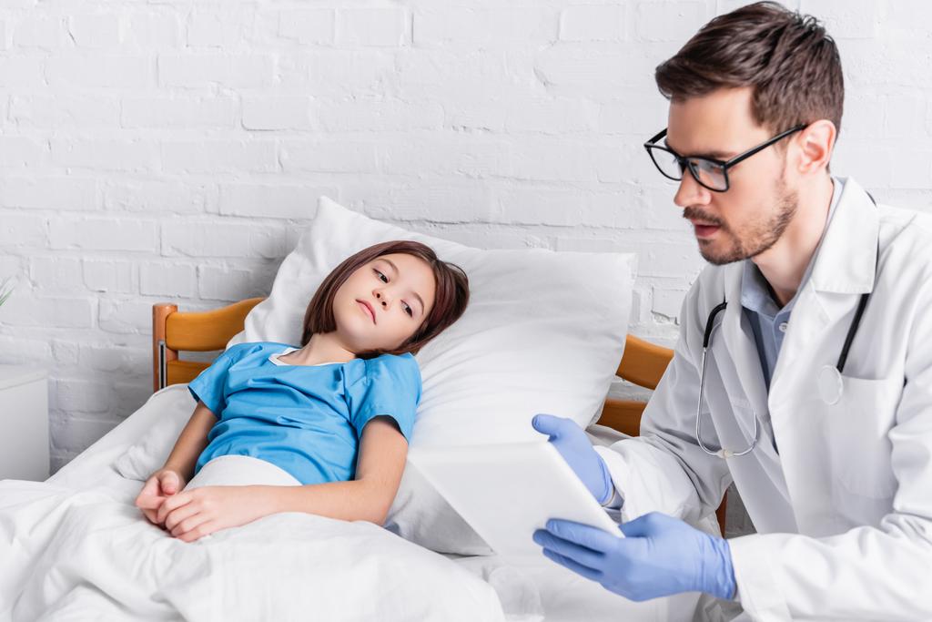 Arzt zeigt auf digitales Tablet, während er mit krankem Kind im Bett spricht - Foto, Bild