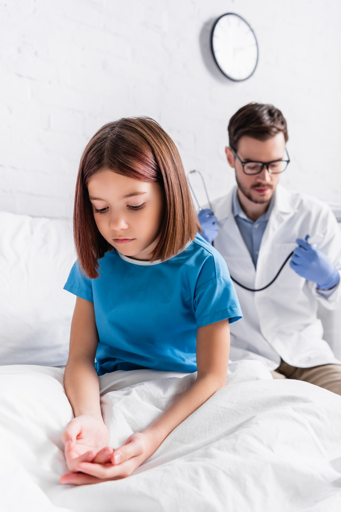 chorej dziewczyny siedzącej na łóżku w klinice w pobliżu pediatry ze stetoskopem na nieostrym tle - Zdjęcie, obraz
