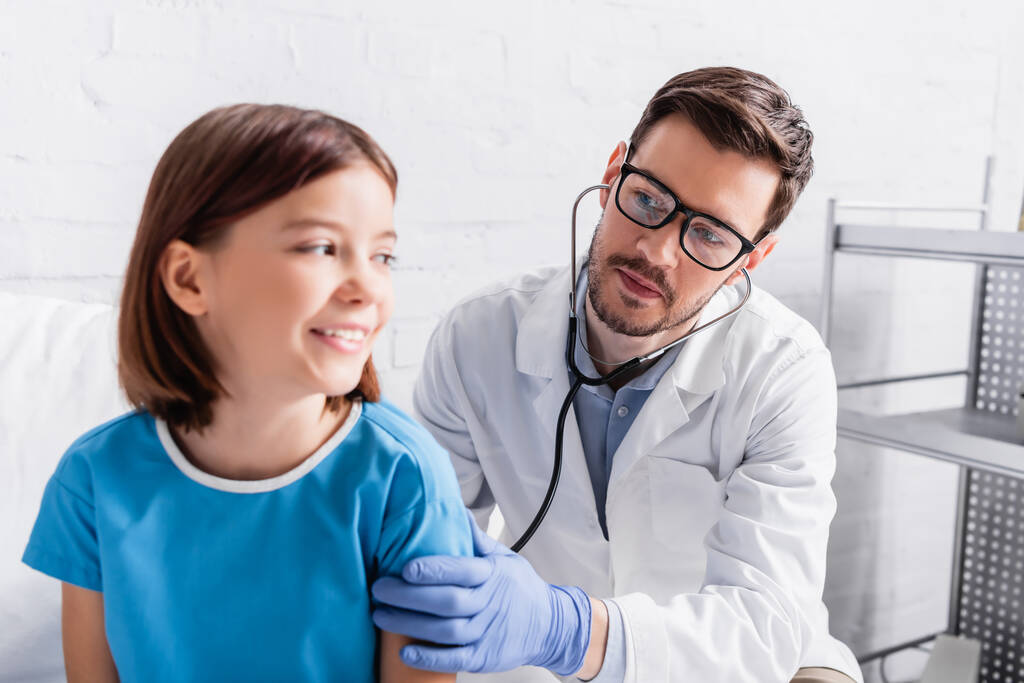 pediatrician examining smiling girl with stethoscope in hospital, blurred foreground - Foto, afbeelding