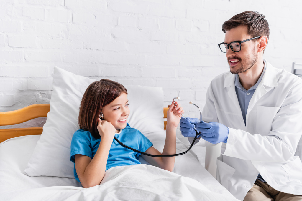 pédiatre joyeux montrant stéthoscope à fille heureuse dans le lit d'hôpital - Photo, image