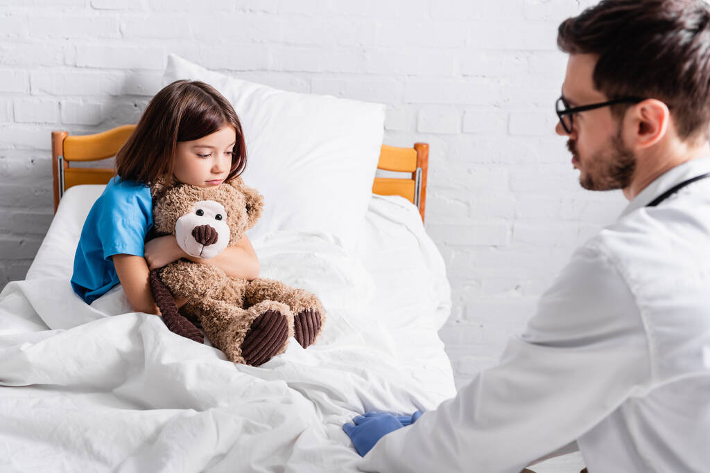 nemocná dívka s medvídkem sedí v posteli v blízkosti lékaře na rozmazané popředí - Fotografie, Obrázek