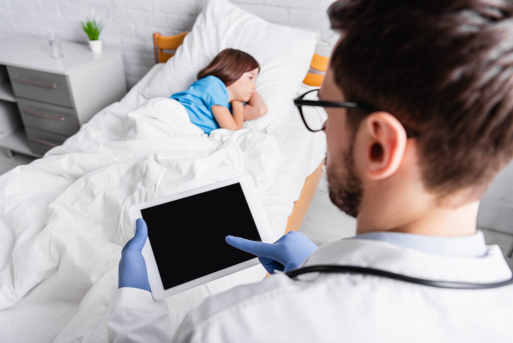 pédiatre à l'aide d'une tablette numérique près d'une fille malade couchée au lit, au premier plan flou - Photo, image