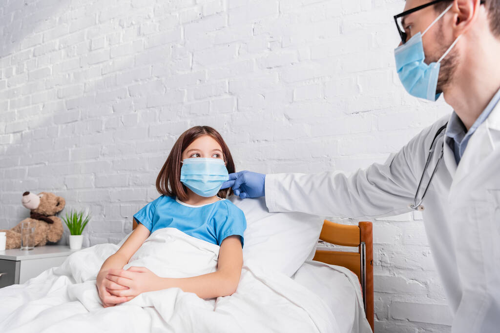Lastenlääkäri lääketieteellinen naamio koskettaa kaulan sairas tyttö istuu sairaalassa sängyssä - Valokuva, kuva