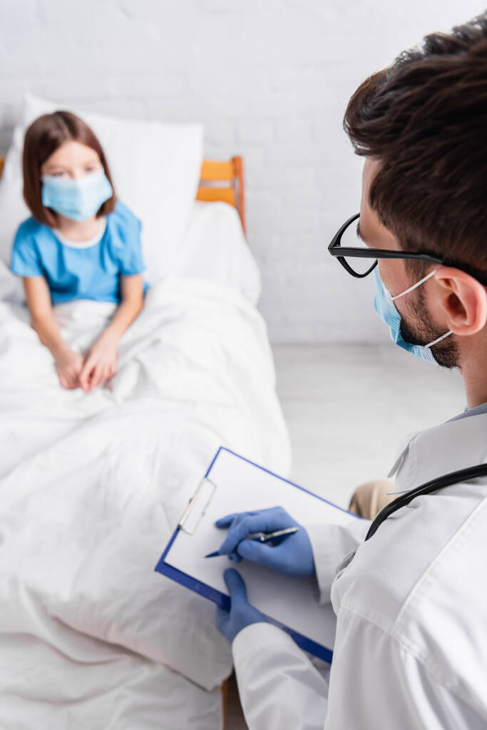pédiatre écriture diagnostic sur presse-papiers près fille malade dans masque médical sur fond flou - Photo, image