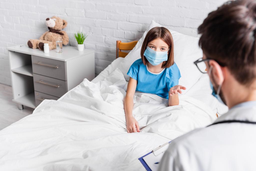 child in medical mask pointing with hand while talking to doctor on blurred foreground - Photo, Image