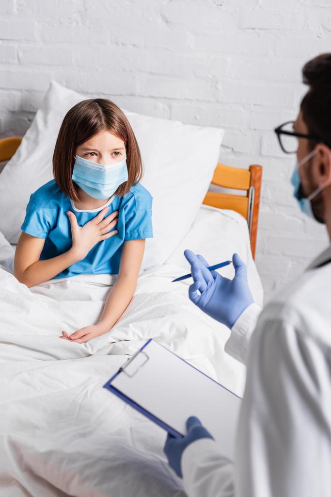 niño con máscara médica tocando el pecho mientras habla con el médico con el portapapeles en primer plano borroso - Foto, imagen