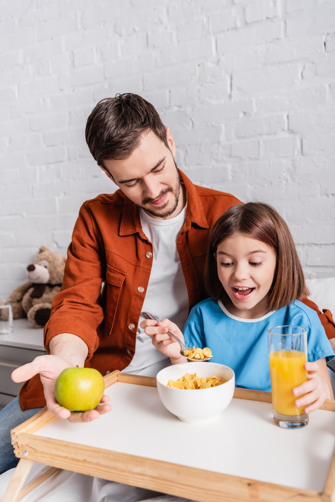 Aufgeregtes Mädchen isst Cornflakes neben Vater, der Apfel im Krankenhaus hält - Foto, Bild