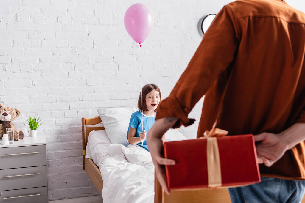 bambino stupito tenendo palloncino festivo vicino a papà con scatola regalo in primo piano sfocato - Foto, immagini