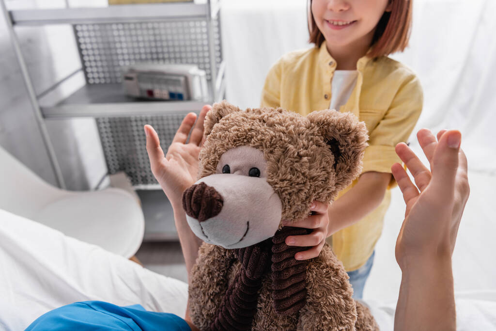 καλλιεργημένη άποψη του χαμογελαστού παιδιού κρατώντας αρκουδάκι κοντά στον πατέρα στο νοσοκομείο - Φωτογραφία, εικόνα