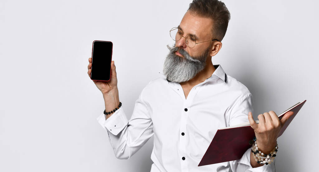 Ein brutaler Mann mit grauem Bart hält ein Buch oder Tagebuch in der Hand und zeigt den Bildschirm seines Mobiltelefons.. - Foto, Bild