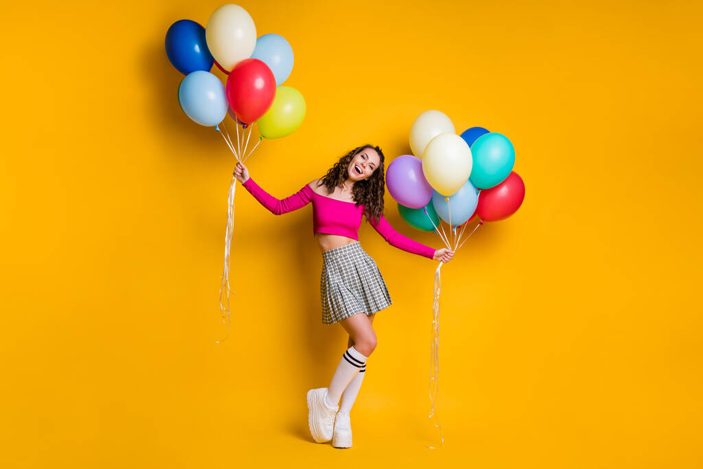 Foto de cuerpo entero del estudiante bastante femenino sosteniendo globos de aire en ambas manos riendo alegremente usando falda a cuadros aislada sobre fondo de color amarillo brillante - Foto, Imagen