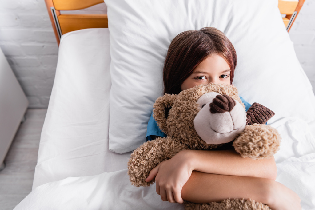vista superior da criança abraçando ursinho de pelúcia enquanto deitado na cama e olhando para a câmera - Foto, Imagem