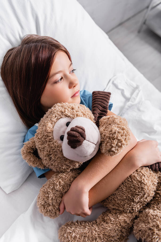 upset, sick girl looking away while embracing teddy bear in hospital bed - Foto, Imagen