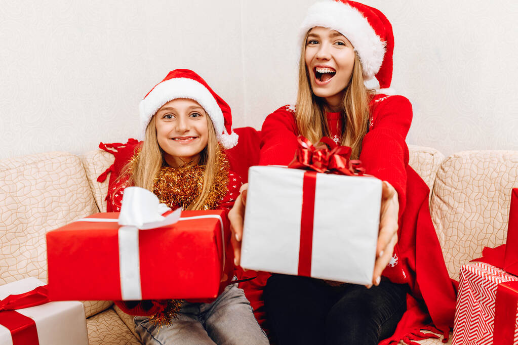 サンタクロースの帽子の幸せな家族、美しい母親と彼女の小さな娘は、贈り物、メリークリスマスとハッピーニューイヤーで家族を愛し、クリスマスプレゼントとソファの上に家に座っています - 写真・画像