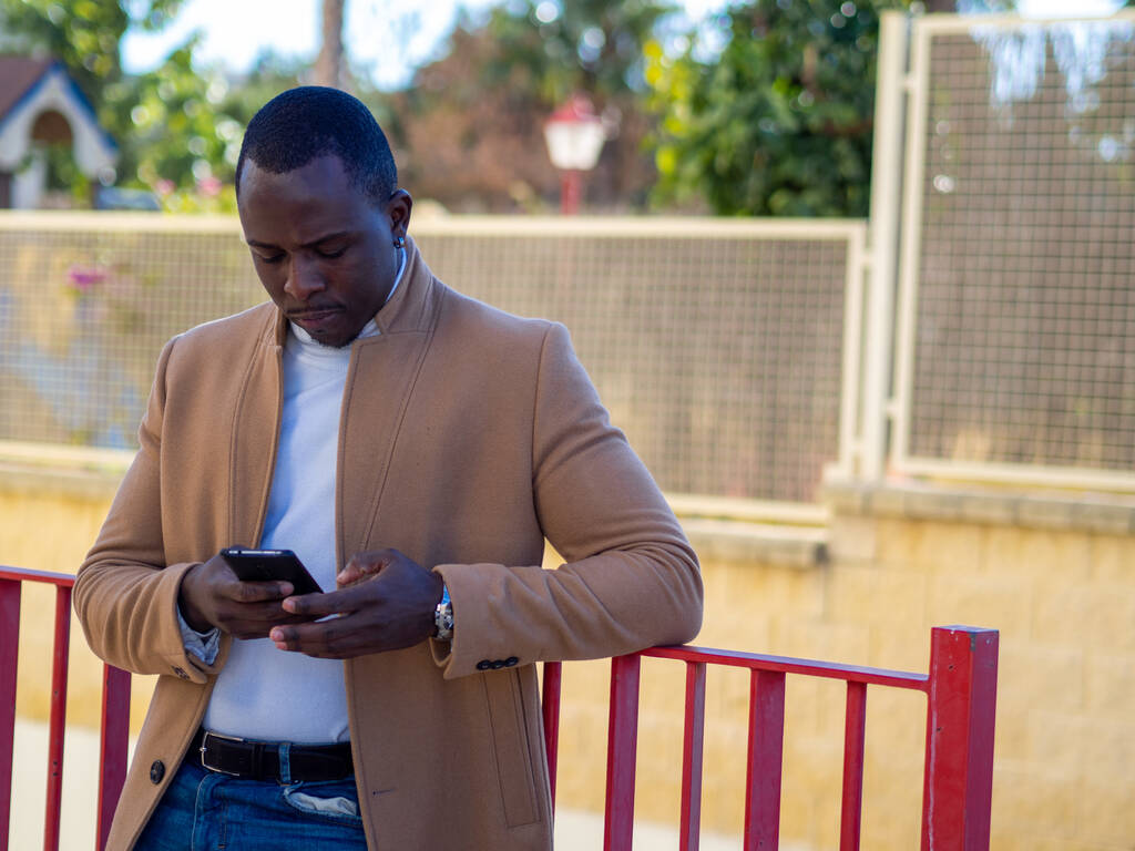 Egy felnőtt, elegáns afrikai férfi sekélyes fókusza, aki a kerítésre támaszkodik és sms-ezik a telefonján. - Fotó, kép
