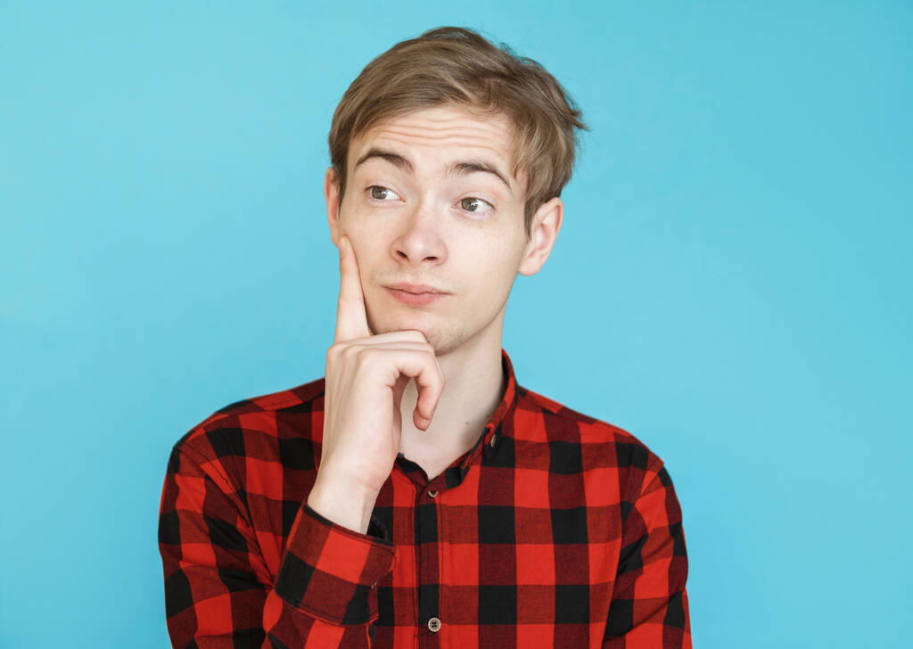 νεαρός άνδρας έφηβος με κόκκινο πουκάμισο σε μπλε φόντο σκέφτεται - Φωτογραφία, εικόνα