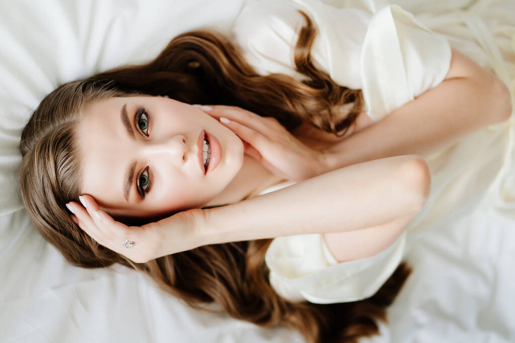 Вид зверху.красива жінка з макіяжем, довге волосся лежить у білому халаті на ліжку
 - Фото, зображення