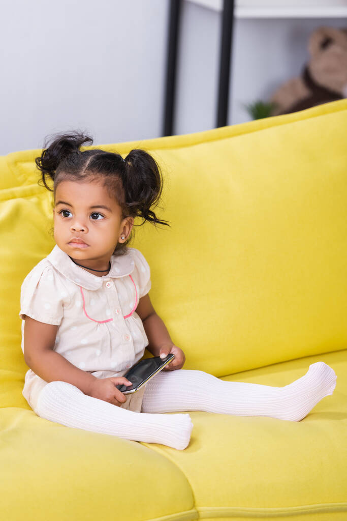 batole africký americký dítě drží smartphone zatímco sedí na pohovce - Fotografie, Obrázek