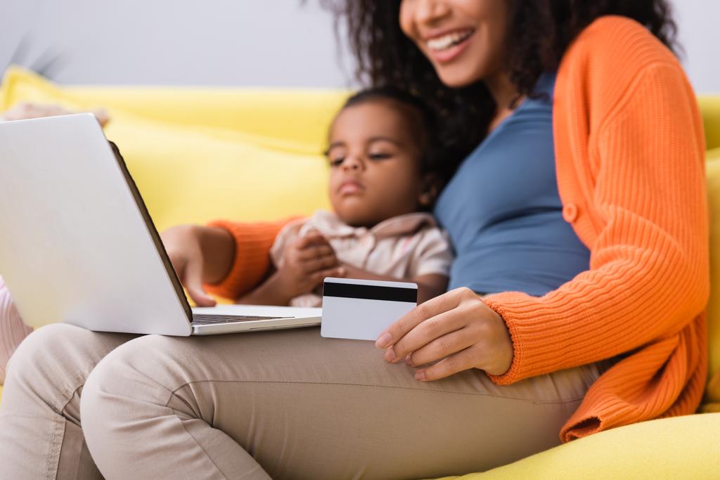heureuse mère afro-américaine tenant la carte de crédit et utilisant un ordinateur portable près de la fille tout-petit dans le salon  - Photo, image