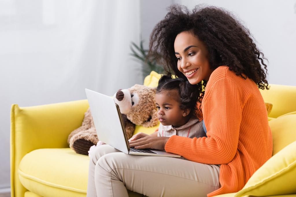 усміхнена афроамериканська мати використовує ноутбук біля дочки малюка у вітальні
 - Фото, зображення